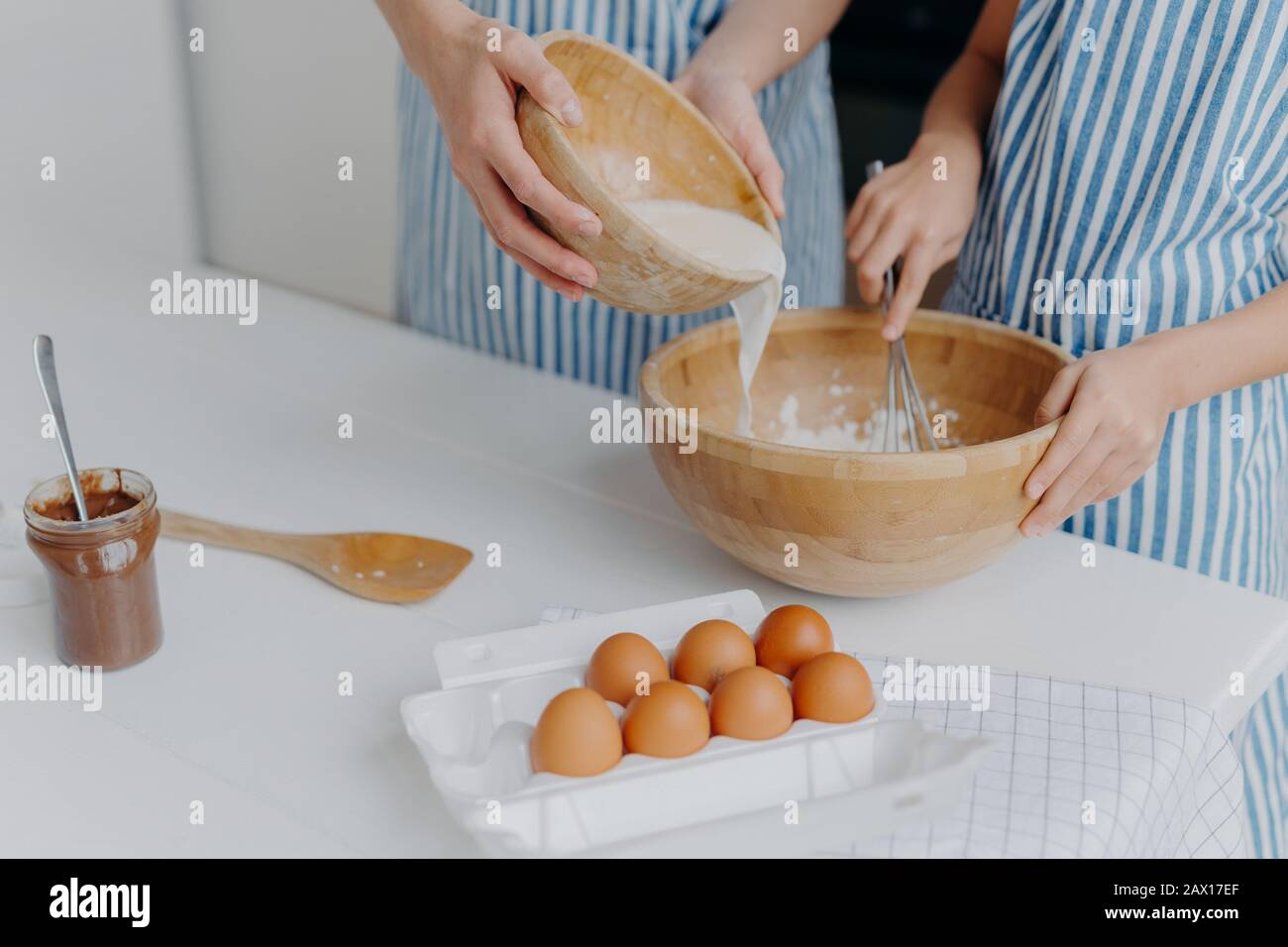 Unerkennbare Mutter und Tochter geben Milch in die Schüssel, machen Teig, hausgemachte Plätzchen tragen Schürzen, stehen auf dem Tisch mit Eiern, Schokolade und Holzspachtel, Stockfoto