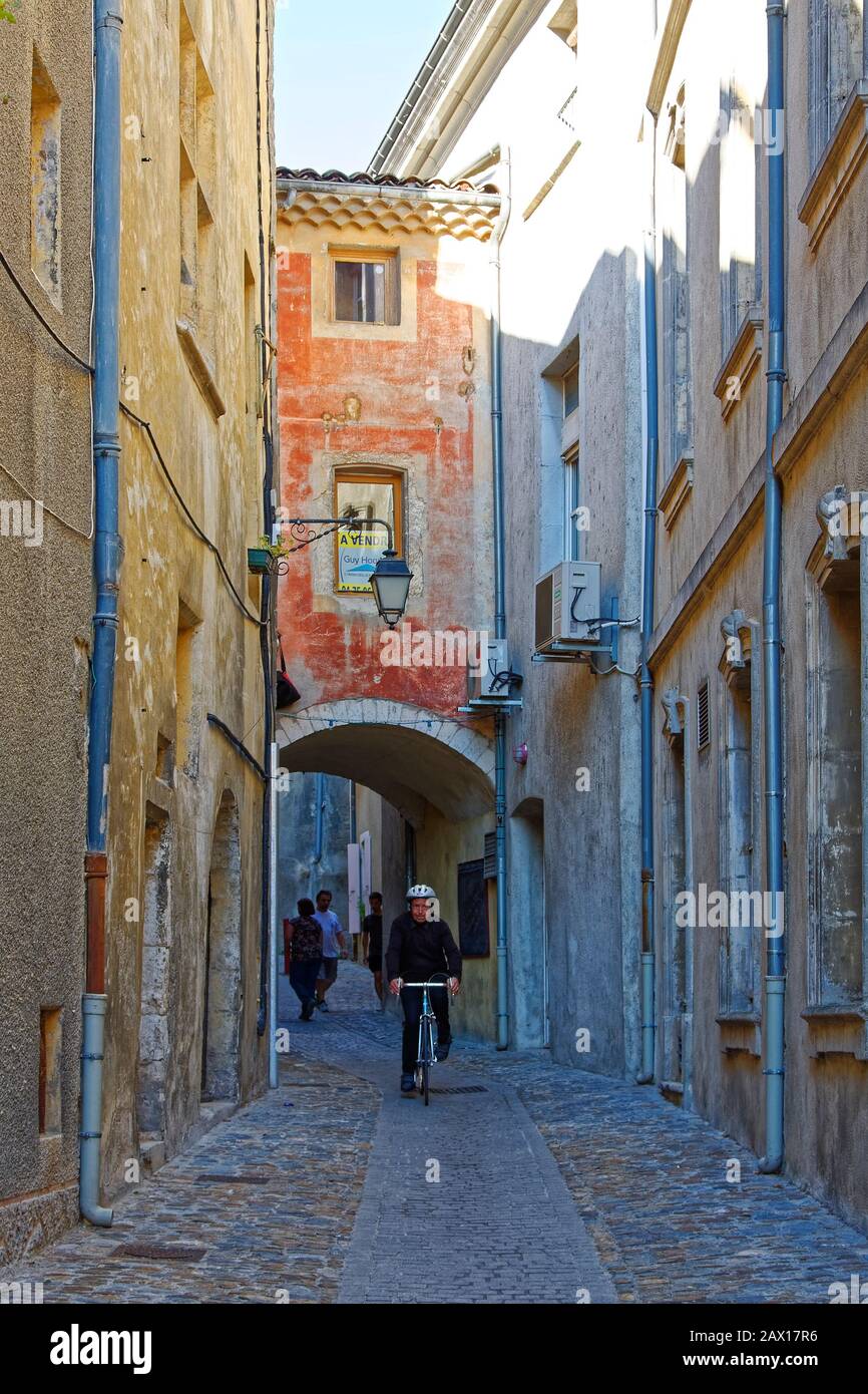 Enge Fußgängerzone, Kopfsteinpflaster, Bogen, alte Gebäude auf der anderen Straßenseite, Radfahrer, Stadtbild, Schatten, Sonne, Viviers, Frankreich; Sommer, vertikal Stockfoto