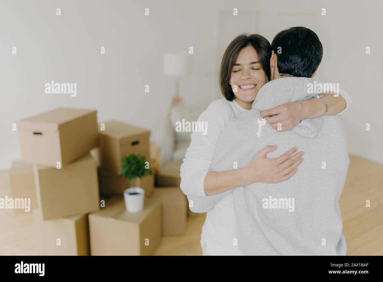 Fröhliche brünette lächelnde Frau umarmt ihren Mann mit Liebe, hält Schlüssel, kauft gemeinsam das erste Haus, kauft neues Grundstück, posiert in unmöbliertem Raumwit Stockfoto