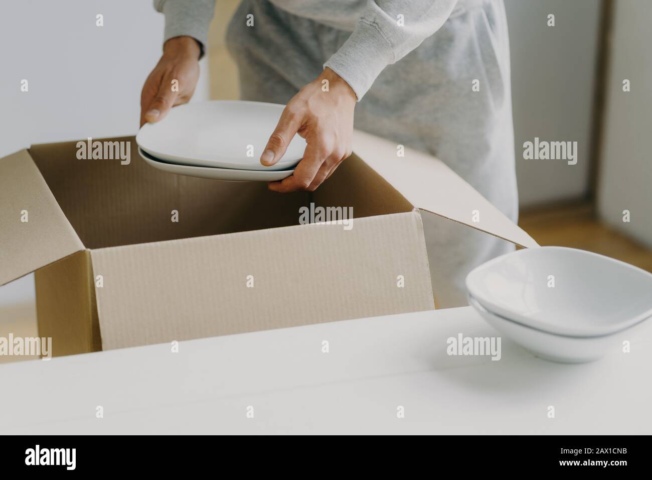 Zugeschnittenes Bild eines unbekannten gesichtslosen beschäftigten Mannes entpackt Kisten mit Geschirr, hält weiße Teller, bewegt sich in neuer Wohnung, tickt in der Küche auf, hat einen beweglichen Tag Stockfoto