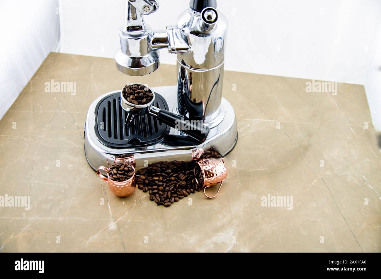 Verschüttete Kaffeebohnen vor einer Espressomaschine, darauf mit einem Hebel aus Bohnen gelegt. Metallbecher in Kupferfarbe. Stockfoto