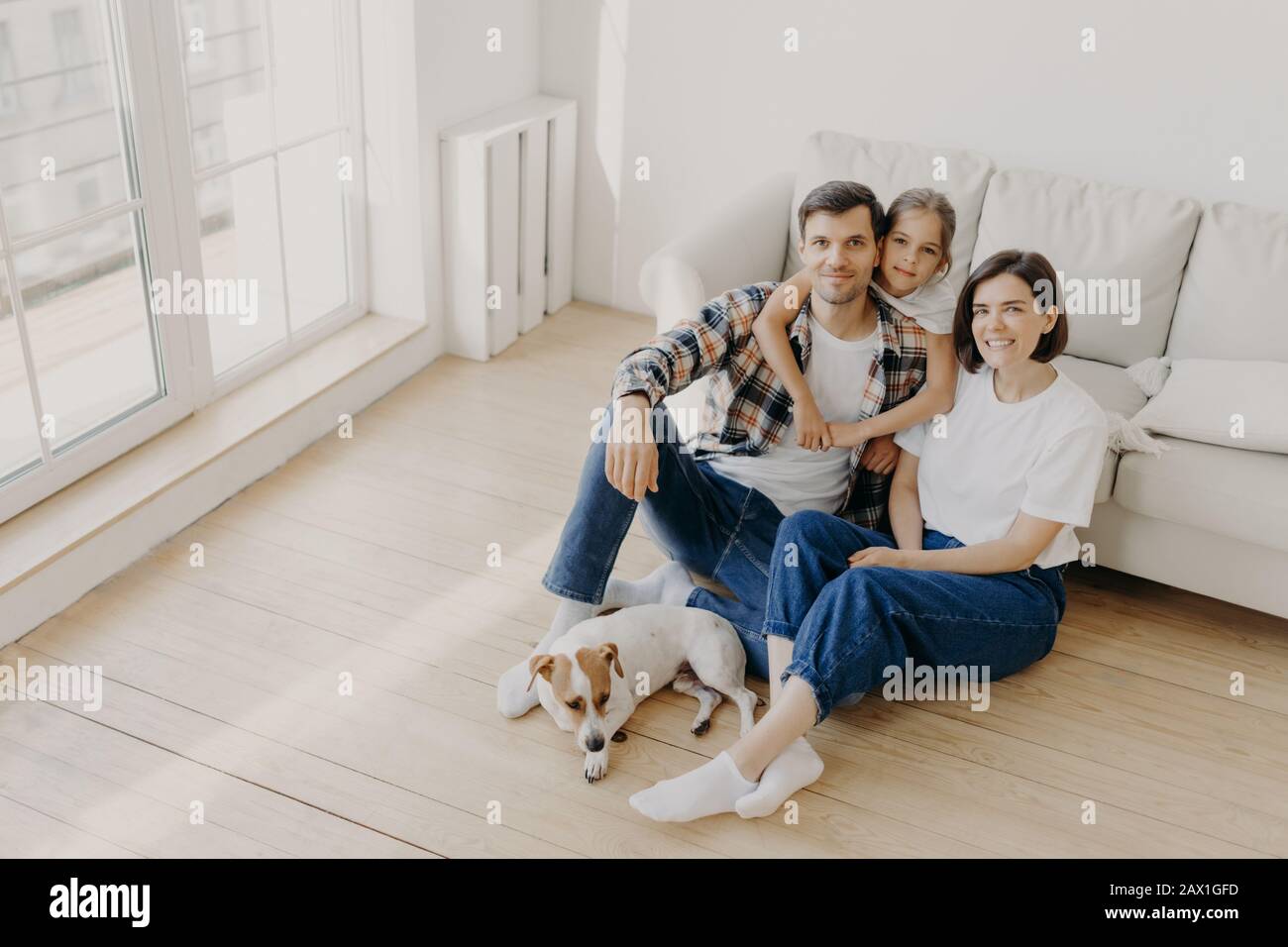 Schöne europäische Familie von Vater, Mutter und ihrer Tochter sitzen auf dem Boden in der Nähe des Sofas in einem geräumigen weißen Raum, Stammhund liegt in der Nähe, haben einen beweglichen Tag, ch Stockfoto
