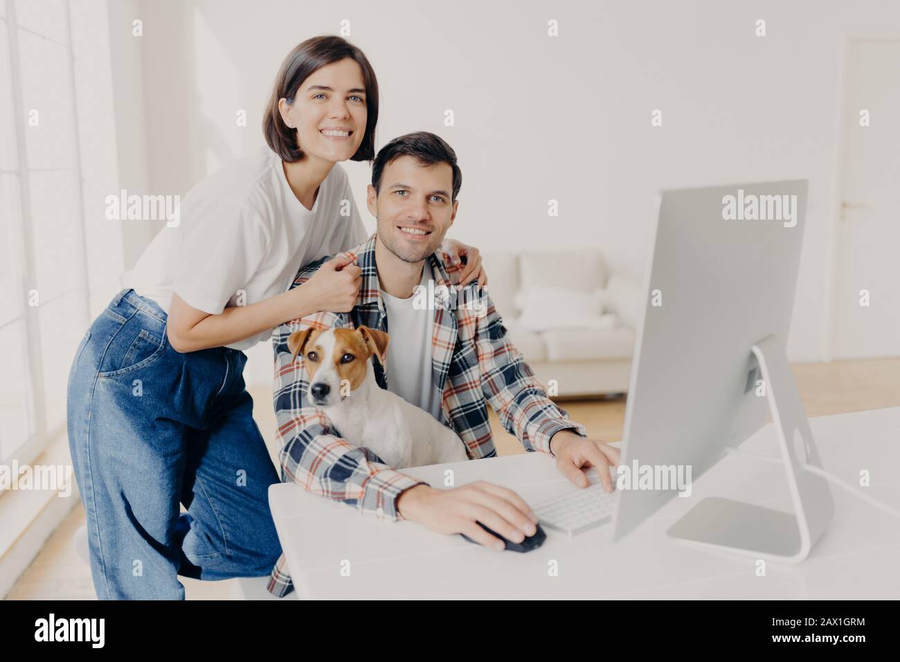 Bild einer lächelnden Frau mit fröhlicher Brunette, die sich an den Schultern der Ehemänner lehnt, die am Computer Tastaturen, im Internet surfen und eine neue Wohnung suchen, um sie auf der Website zu kaufen, Stockfoto