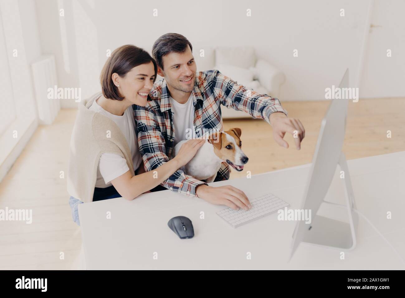 Überfreute Frau und Mann haben fröhliche Blicke, schauen sich Online-Filme zuhause an, spielen mit Punkt, sitzen am weißen Tisch, suchen Informationen über wifi, gekleidet Stockfoto