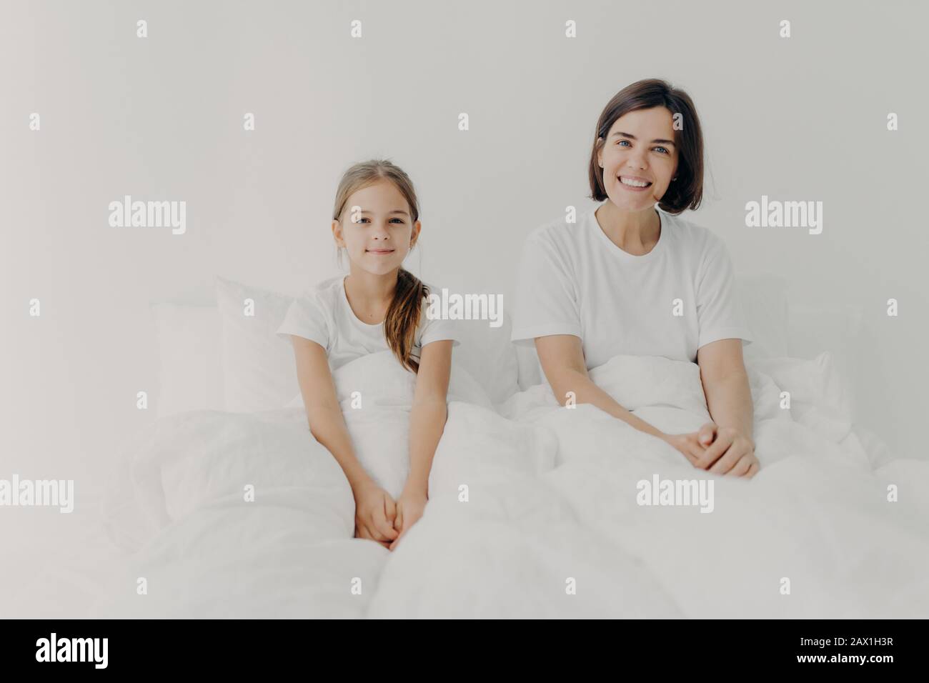 Morgenzeit und Erweckungskonzept. Fröhliche lächelnde Mutter und Tochter sitzen in einem komfortablen Bett, in weißen T-Shirts gekleidet, wach mit positiver Stimmung, gla Stockfoto