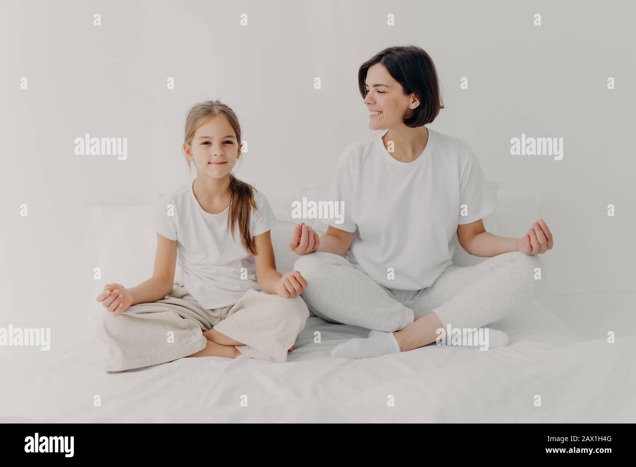 Horizontaler Schuss von fröhlicher entspannter Brunette Mutter und Tochter posieren in lotus-pose, versuchen Sie sich zu entspannen, halten Sie die Beine gekreuzt, gekleidet in lässiger weißer Kleidung, ich Stockfoto