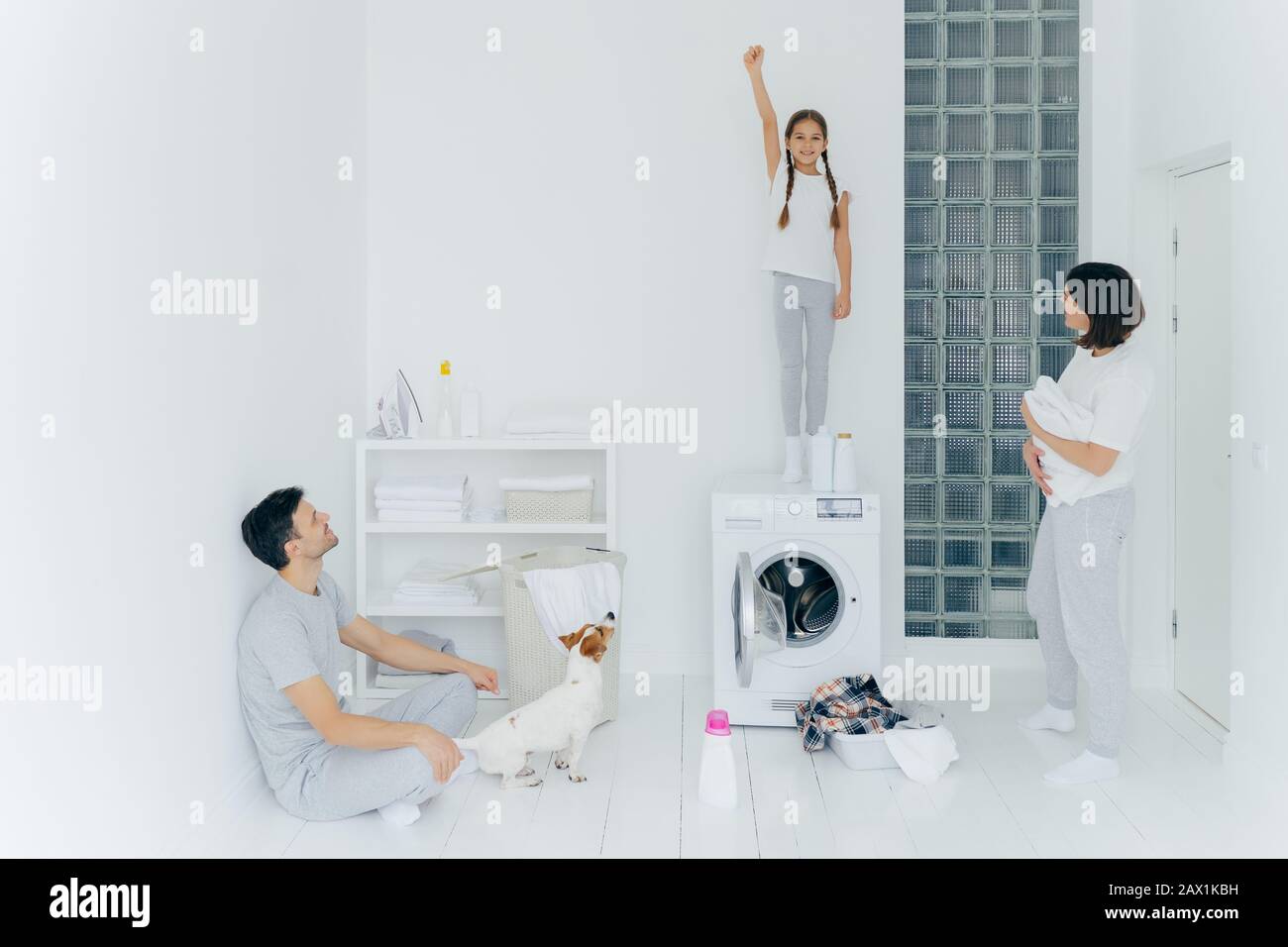 Foto des fröhlichen Mädchens steht auf der Waschmaschine, hebt Arm mit geklenkelter Faust, hilft Eltern, Kleidung zu waschen, Mutter, Vater und Hund schauen auf Kind. Fa Stockfoto