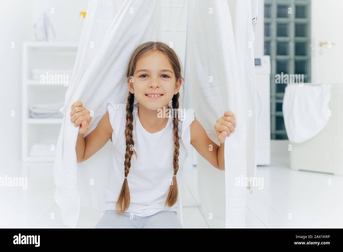 Glückliches europäisches Mädchen mit zwei Zöpfen, posiert in der Nähe von Kleiderpferd zwischen weißem trocknendem Leinen, posiert im Waschraum vor verschwommenem Hintergrund, weißer Colo Stockfoto