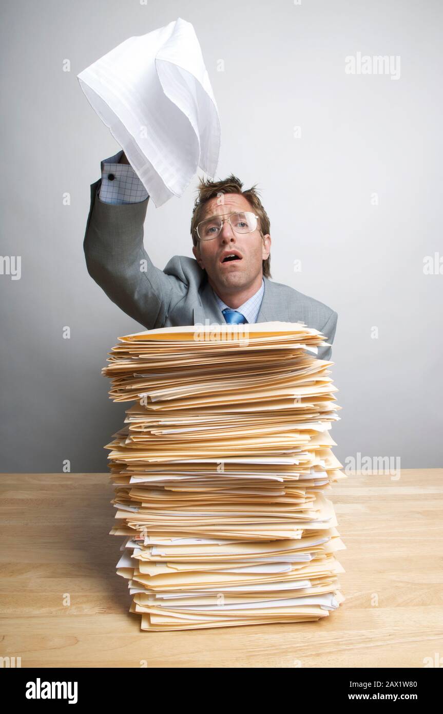 Verzweifelter Büroangestellter, der an seinem Schreibtisch sitzt und eine weiße Fahne der Kapitulation über einem großen Stapel von Papierkram schwenkt Stockfoto