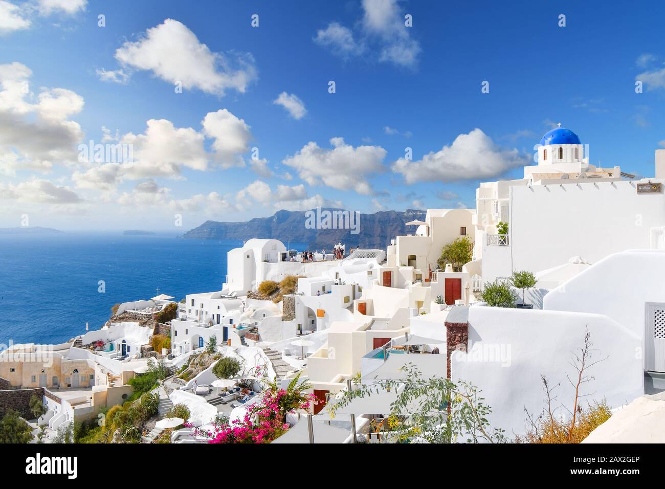 Oia, Santorini, Griechenland im Sommer. Weiß getünchte Häuser und blaue Kuppelkirche unterstreichen die Insel, während Touristen einen Blick auf die Terrasse des Resorts auf das Meer genießen. Stockfoto