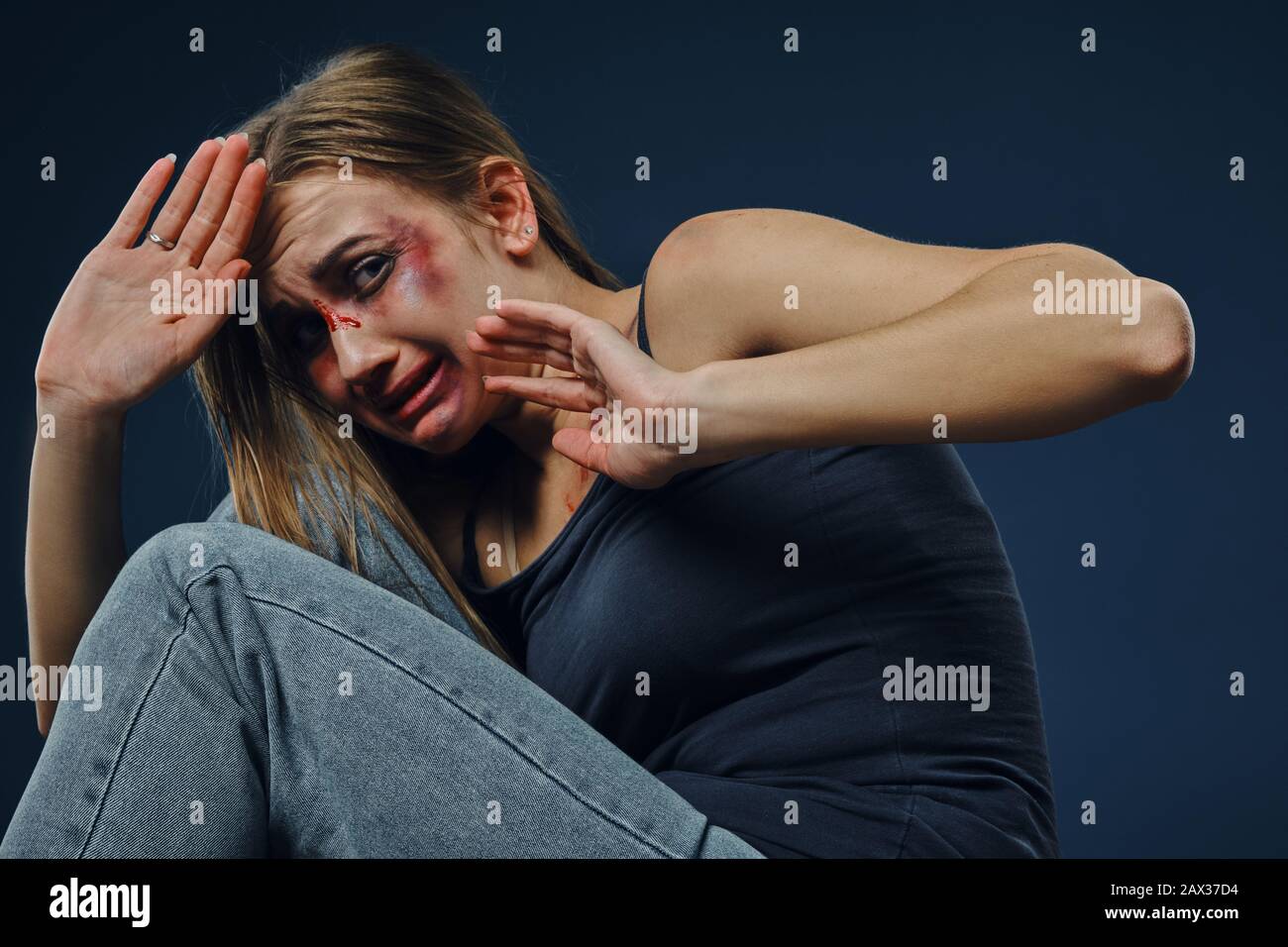 Trauerte blonde Frau in schwarzem Unterhemd und Jeans. Blutendes Gesicht mit Prellungen bedeckt, hinter den Händen versteckt, seitlich vor blauem Hintergrund sitzend. Stockfoto