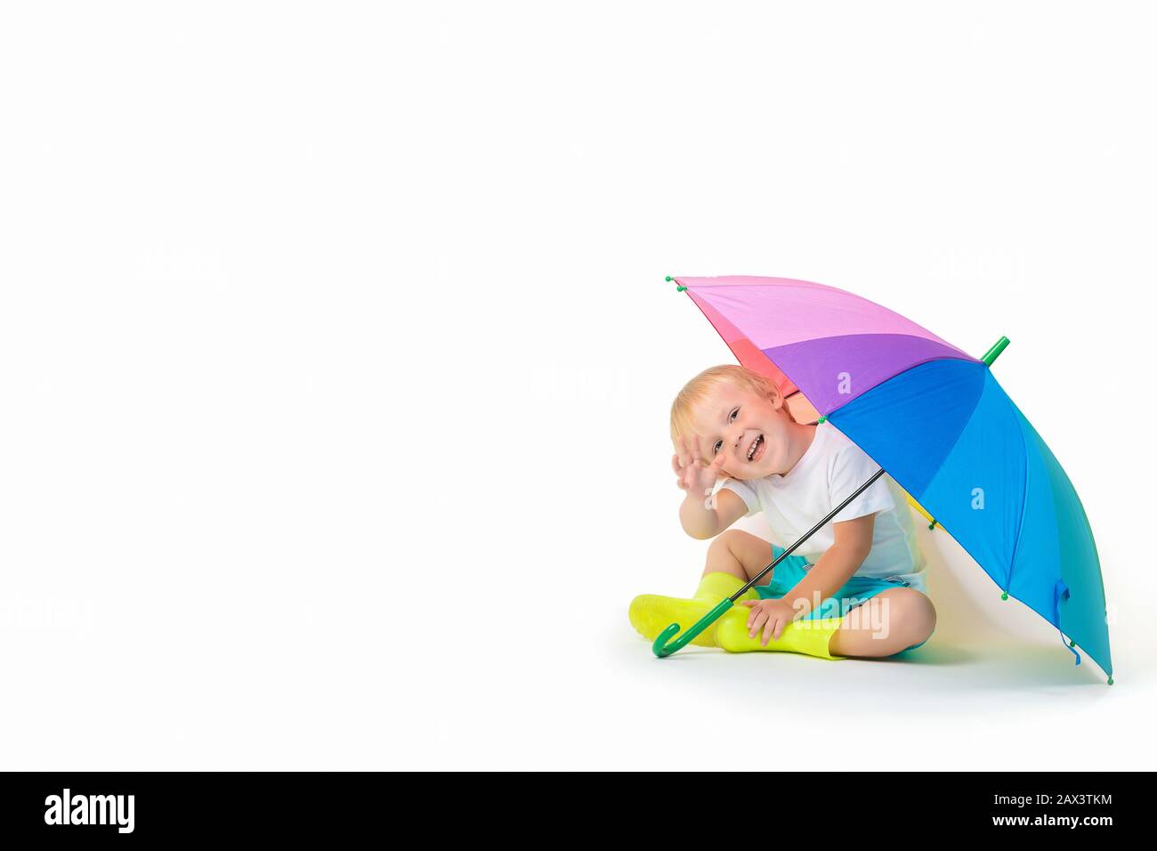 Ein blondes Kind kaukasisch sitzt unter einem regenbogenhellen Regenschirm in Gummistiefeln und Wellen seine Hand auf einem weißen Hintergrund. Konzept über Puddel, Sicherheit Stockfoto
