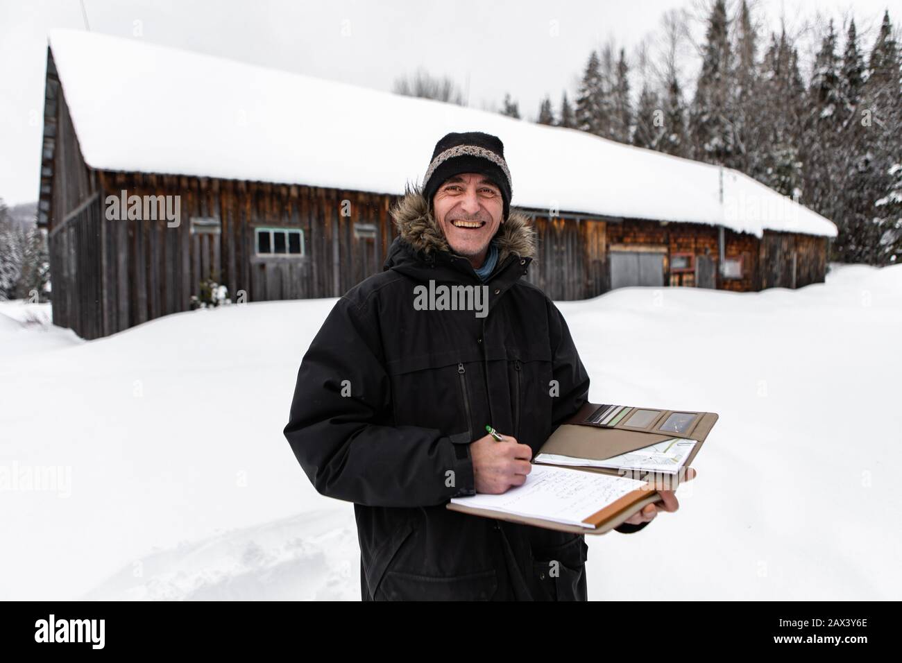 Hausinspektor mittleren Alters, der bei einer Inspektion Notizen machte. Lächelnder Mann mit Winterkleidung steht draußen im Schnee, Bauernhaushintergrund in Fachwerk Stockfoto