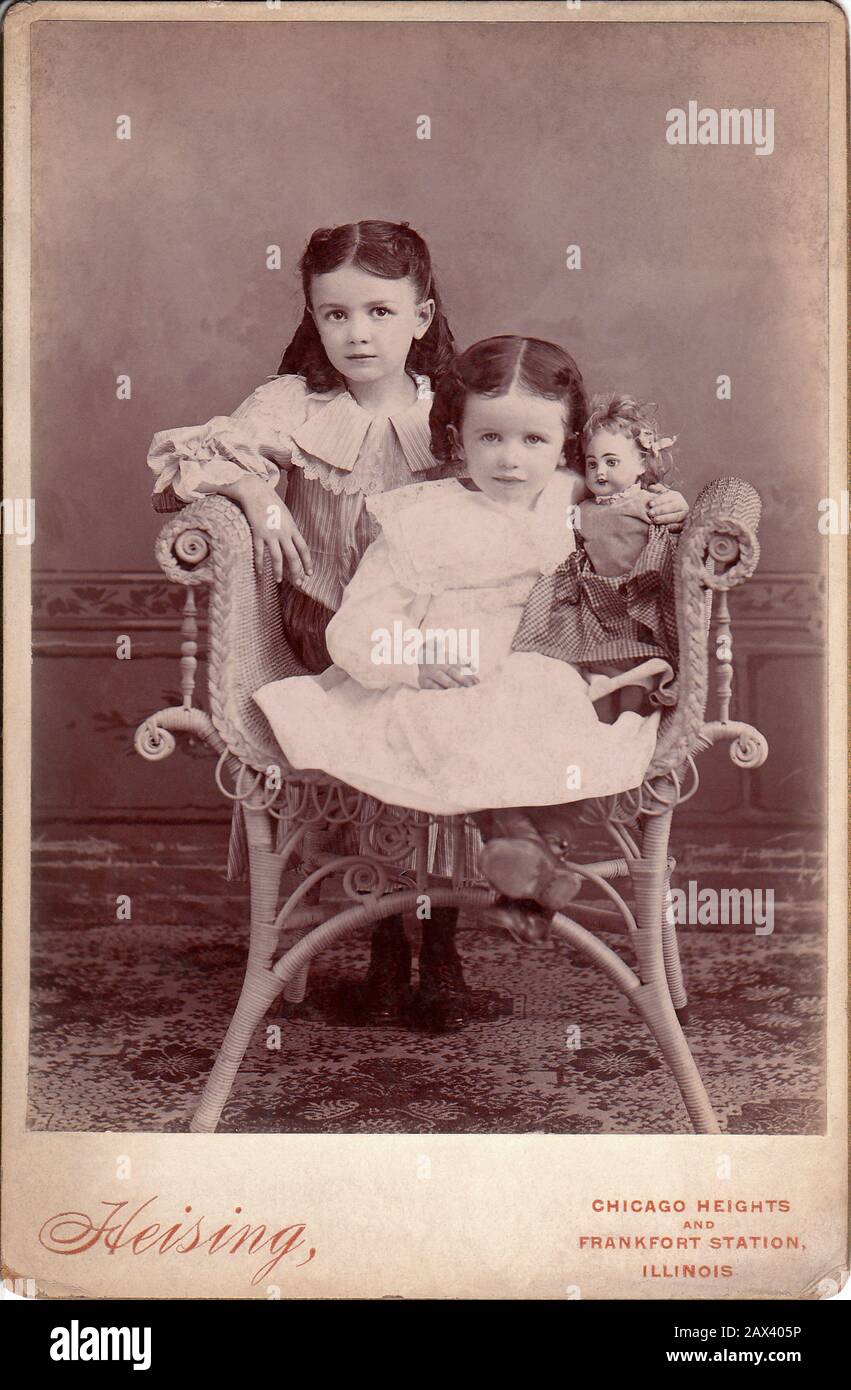 Ca, CHICAGO, USA: Kleine Mädchen mit Puppe . Foto von Heising , Chicago ILLINOIS - FOTO STORICHE - HISTORIENFOTOS - BAMBINA - KIND - BAMBINO - BAMBINI - BAMBINE - KINDER - TENEREZZA - ZÄRTLICH - KINDER - BAMBINA - BABY - OTTOCENTO - BELLE EPOQUE - MODE - MODA-INFANTILE - BABY - BEBÉ - ROSA KLEID - KINDHEIT - INFANZIA - Bambola - Bambol - Spielzeug - Giocattolo - Giocattoli - gioco - Spiel - poltrona di vimini - Rattan - pizzo - Spitze - Sorella - sorelle - Schwestern ---- Archivio GBB Stockfoto