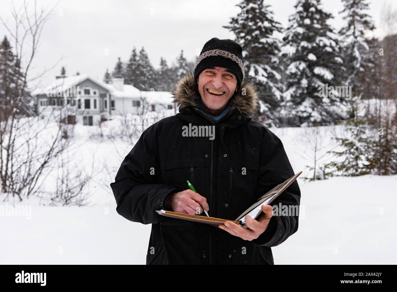 Selektiver Fokus auf Mittelalter-Hausinspektor, der während einer Inspektion Notizen auf einem Notizbuch hält. Ein lächelnder Mann steht im Winter im Schnee Stockfoto
