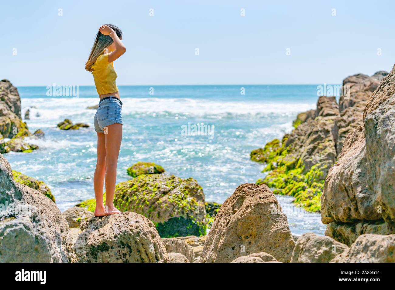 Mädchen in gelbem Oberteil an der felsigen Küste an der Küste am Mount Maunganui, New Zraland. Stockfoto