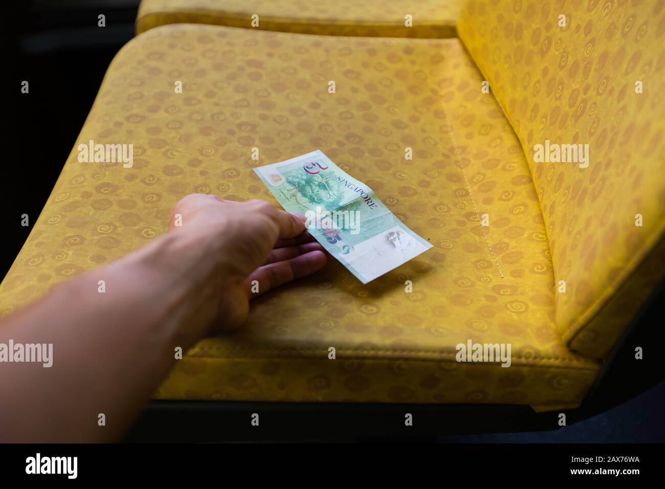 Eine Hand streckt sich aus, um die Singapur 5-Dollar-Banknote links auf dem gelben Sitz im Bus zu nehmen. Stockfoto