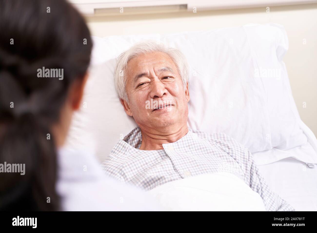 Asiatischer alter Mann, der im Bett liegt und mit junger Ärztin in der Krankenstation spricht Stockfoto