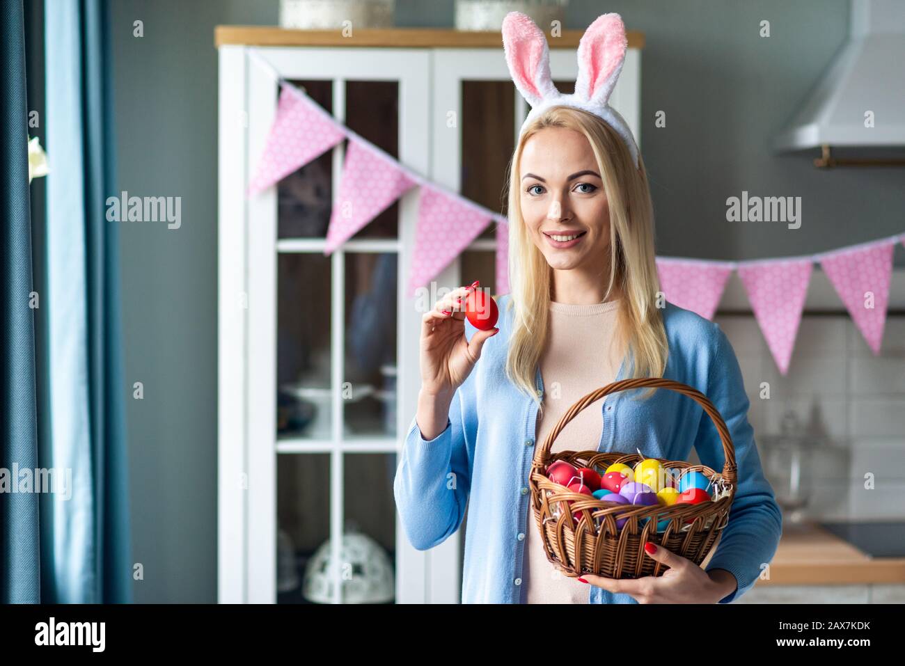 Porträt einer niedlichen Frau, die in Hunneneiern und im stehen trägt. Sie hält einen Korb mit bunten Ostereiern und zeigt einen davon Stockfoto