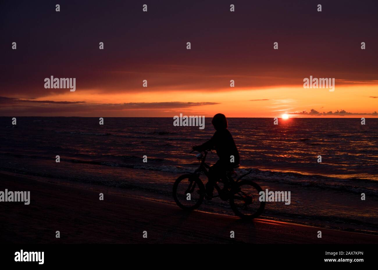 Ein Radfahrer reitet auf dem Meer mit einem Hintergrund der Sonne, die in die dunkle Wolke des Meeres darüber untergeht, und Lichtreflexionen auf kleinen Wellen Stockfoto