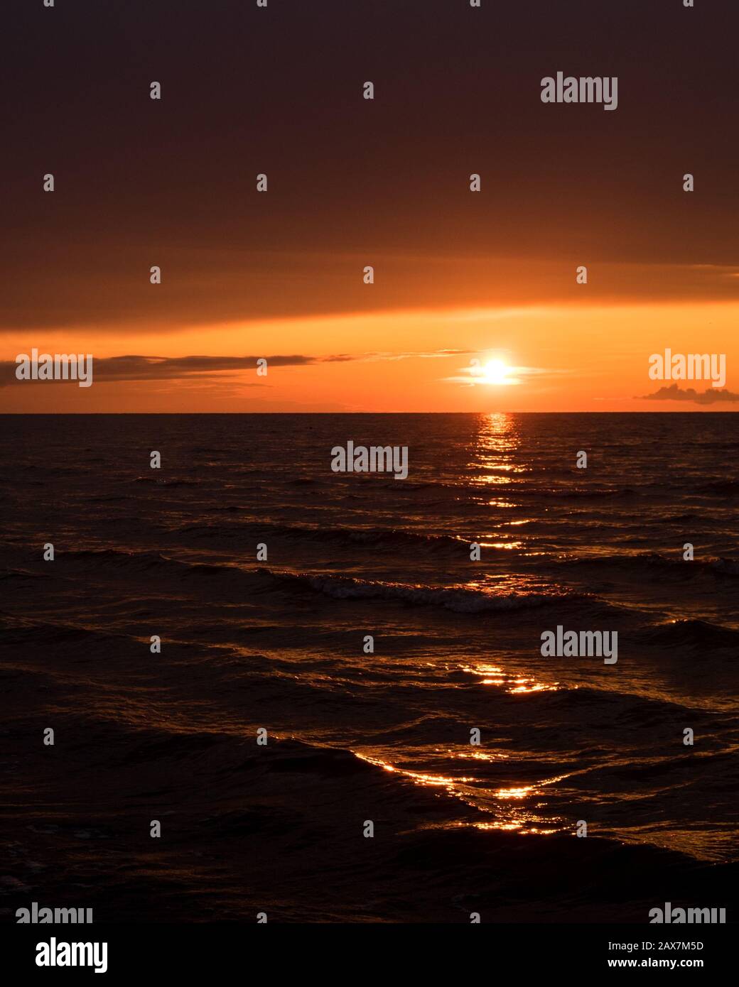 Sonne geht ins Meer mit dunkler Wolke darüber und Lichtreflexionen auf kleinen Wellen senkrecht Stockfoto