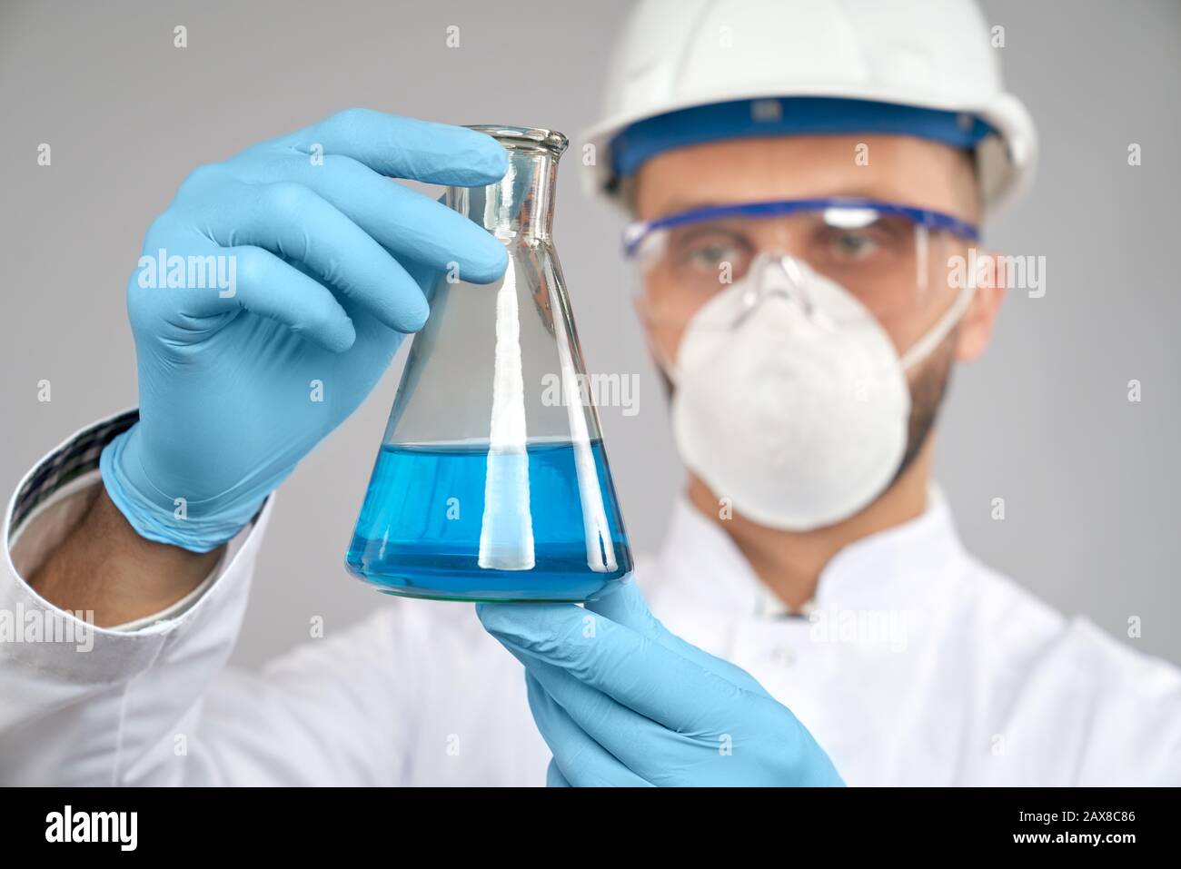 Nahaufnahme des jungen Chemikers in Brille, Helm und Maske, der im Labor experimentiert, isoliert auf Grau. Selektiver Fokus von Kolben mit blauer Flüssigkeit in der Hand des Wissenschaftlers in Handschuhen und weißem Labormantel. Stockfoto
