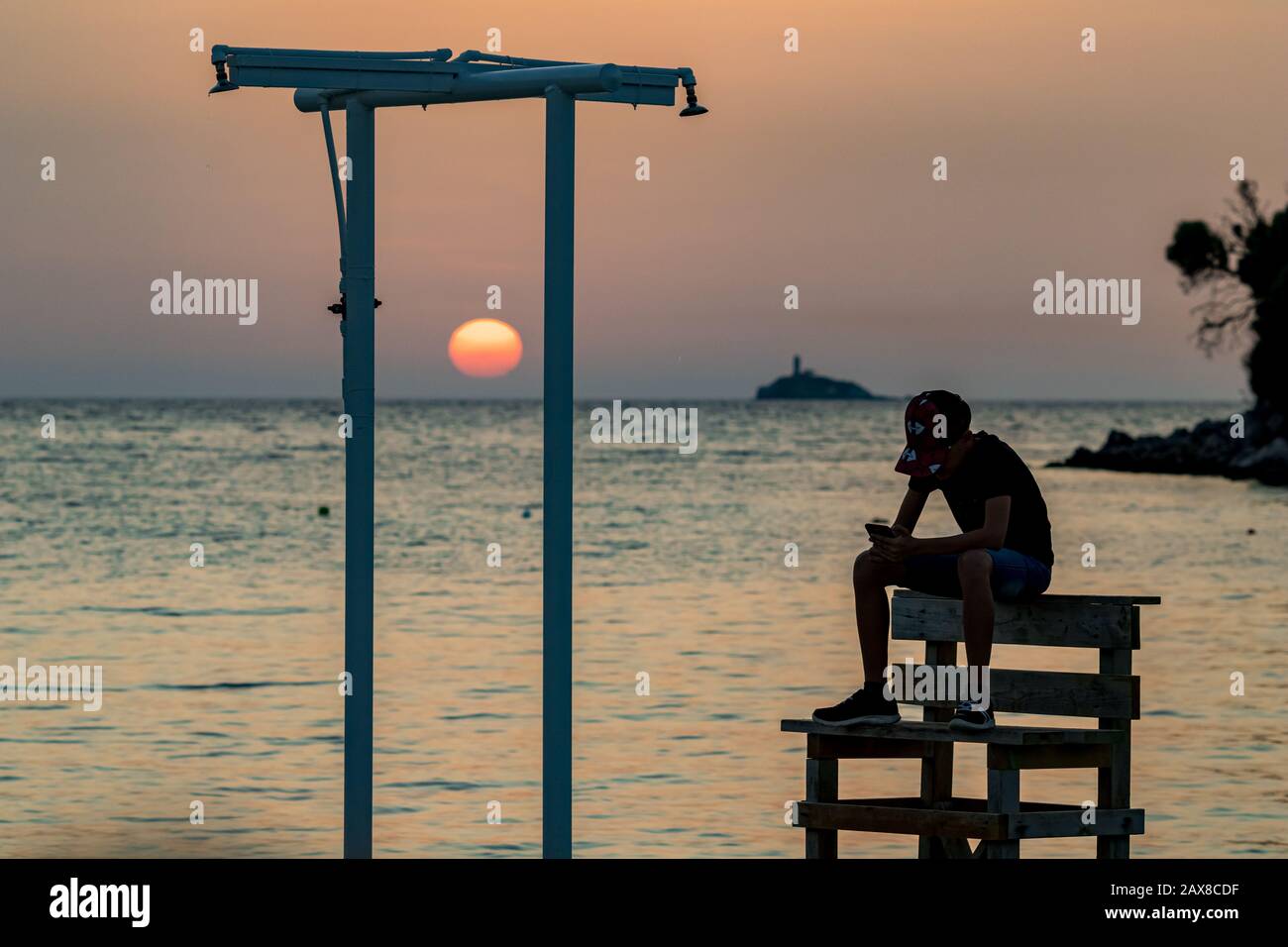 Schöne männliche Silhouette eines nicht erkennbaren schlanken Jungen mit einem Telefonspiel, das bei Sonnenuntergang vor dem goldenen Ionischen Meerwasser in der Nähe des Strandes auf der Bank sitzt. Abenddämmerung von Ksamil aus, Albanien, Frühling Stockfoto