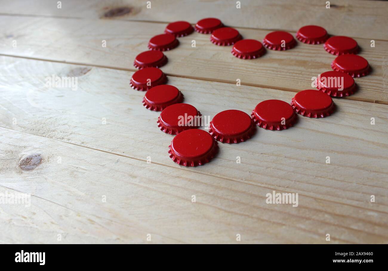 Ein rotes Liebesherz aus Bierflaschenoberteilen, von der Seite auf einem rustikalen Holztisch betrachtet. Bier trinkt Valentinstag Konzept. Selektiver Fokus. Stockfoto