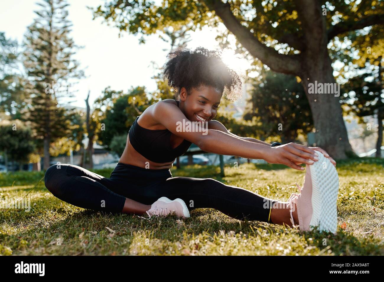 Lächelndes Porträt einer sportiv sitzenden afroamerikanischen jungen Frau auf dem Rasen, die ihre Beine im Park streckt - fröhliche junge schwarze Frau, die sich aufwärmt Stockfoto