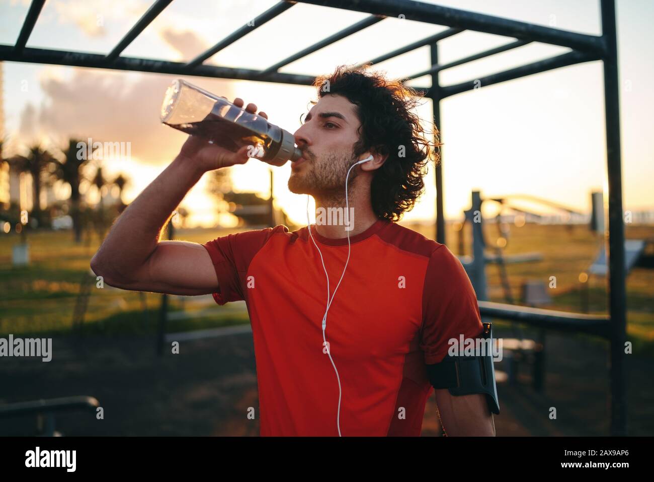 Sportlicher junger kaukasischer Mann, der Musik auf Ohrhörern hört, die Wasser aus einer transparenten, wiederverwendbaren Flasche im Sportpark im Freien trinken Stockfoto