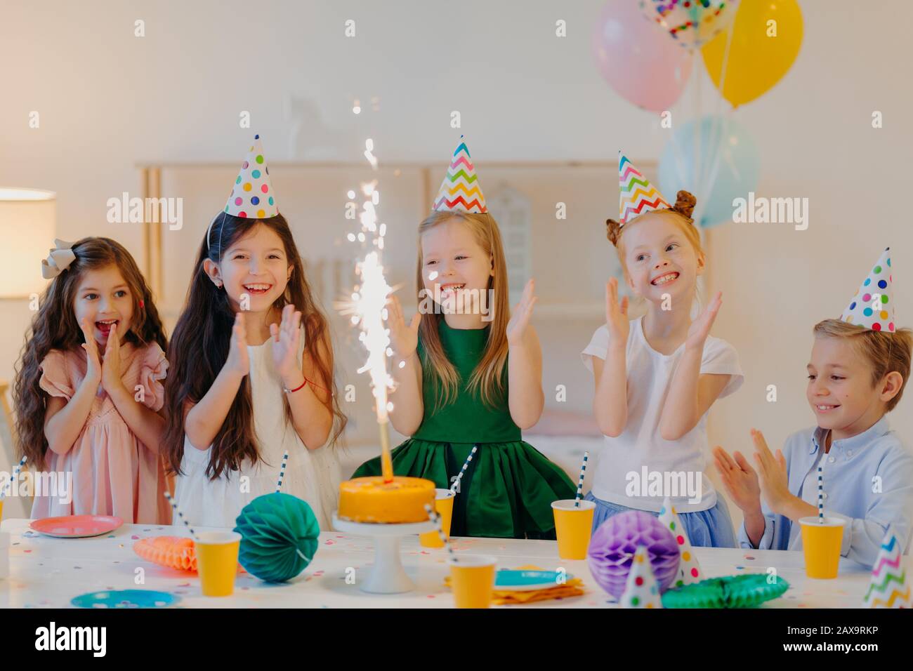 Feiertag- und Festtagskonzept. Glückliche fünf kleine Kinder klatschen die Hände, schauen auf Glitzern von Kuchen, feiern Geburtstag von Freunden, sammeln sich in der Nähe eines großen Tisches, Stockfoto