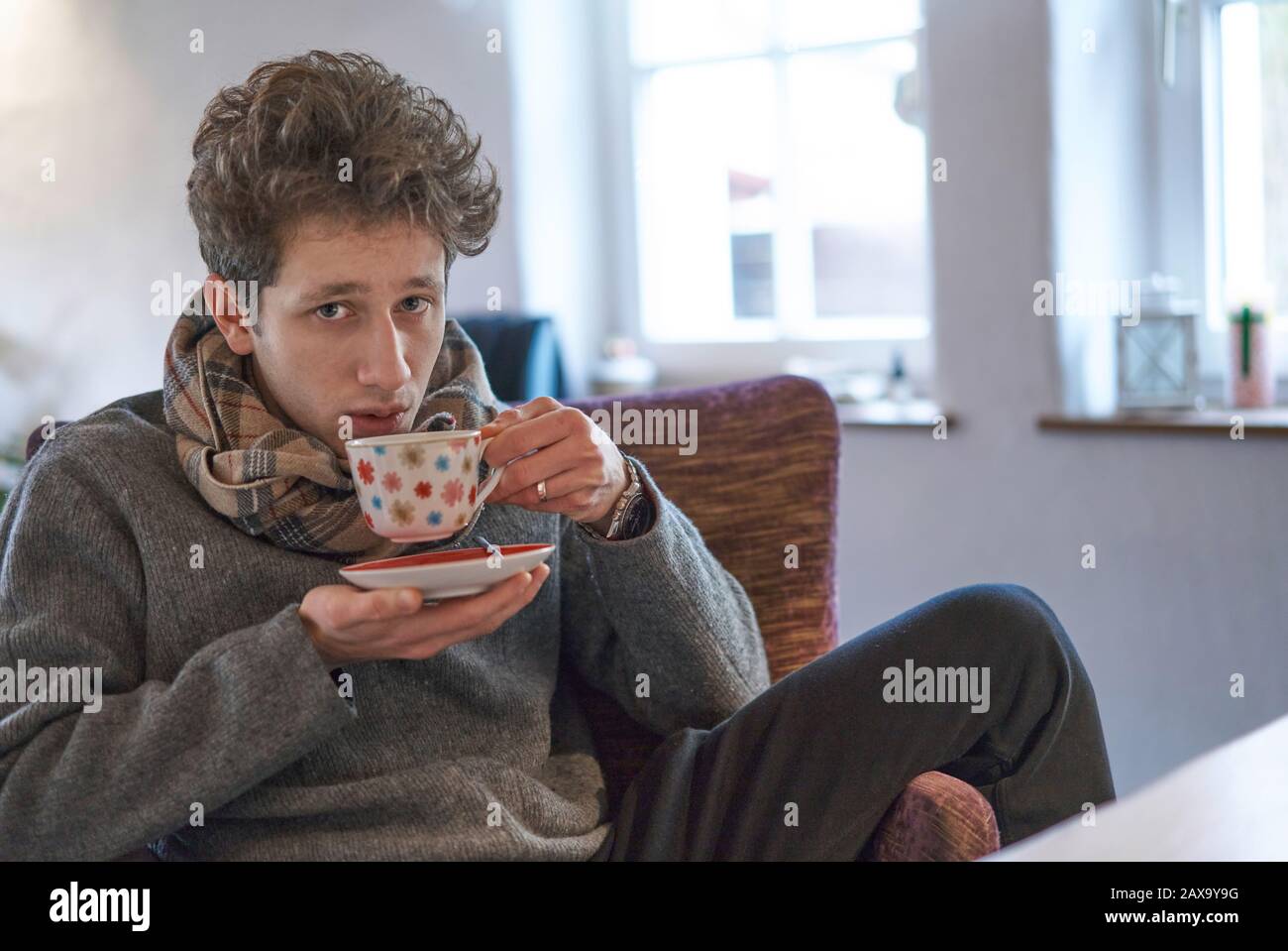 Der junge Mann sitzt entspannt zu Hause auf dem Sessel und trinkt eine Tasse Tee, der etwas krank aussieht Stockfoto