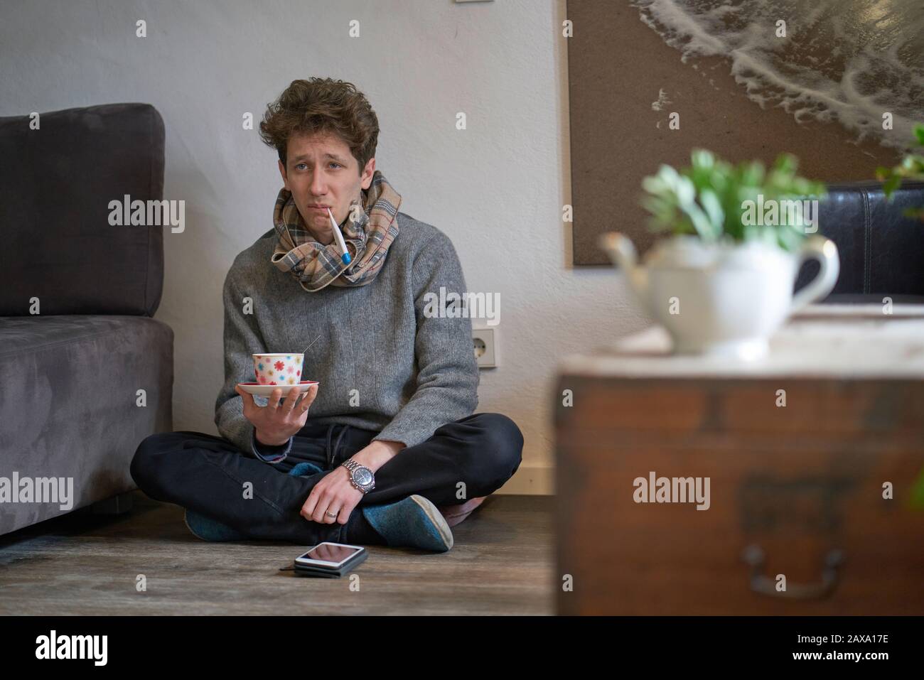 Junger depressiv aussehender Mann mit einem kalten Sitzen auf dem Boden in seiner Wohnung mit einem Thermometer im Mund und einer Tasse Tee in der Hand Stockfoto