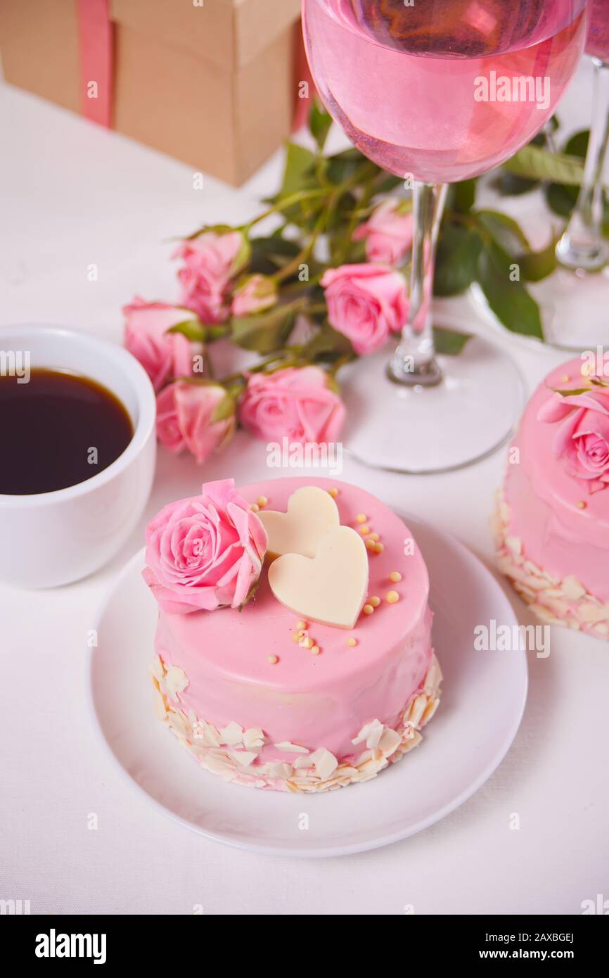 Minikuchen mit rosafarbener Glasur, schönen Rosen, Tasse Kaffee, Glas rosafarbenen Wein auf dem weißen Tisch Stockfoto