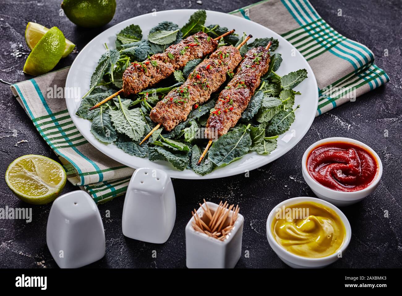 Lammkebab auf Spießen mit frischen, von Grüns gesprinklerten Kaleblättern und gehackten chiles auf einem weißen Teller mit Ketchup, Senf und Kalkkeilen auf einem serviert Stockfoto