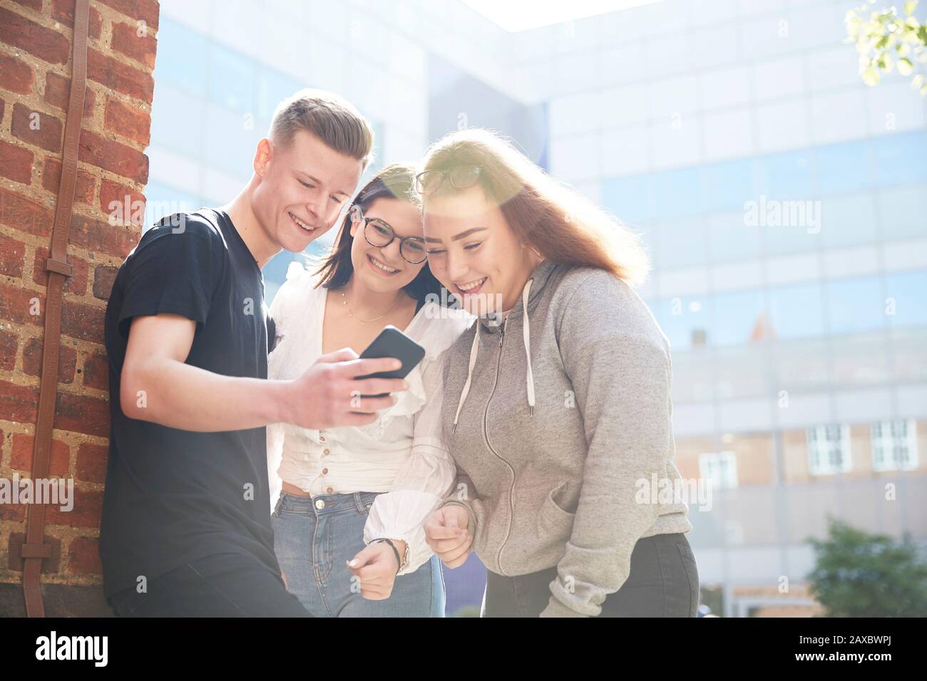 Teenager-Freunde, die ein Smartphone außerhalb des sonnigen Schulgebäudes verwenden Stockfoto