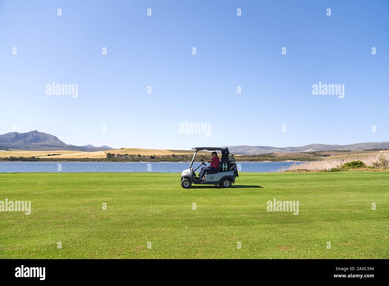 Männlicher Golfer, der auf dem sonnigen Golfplatz auf dem See entlang eine Golfkarre fährt Stockfoto