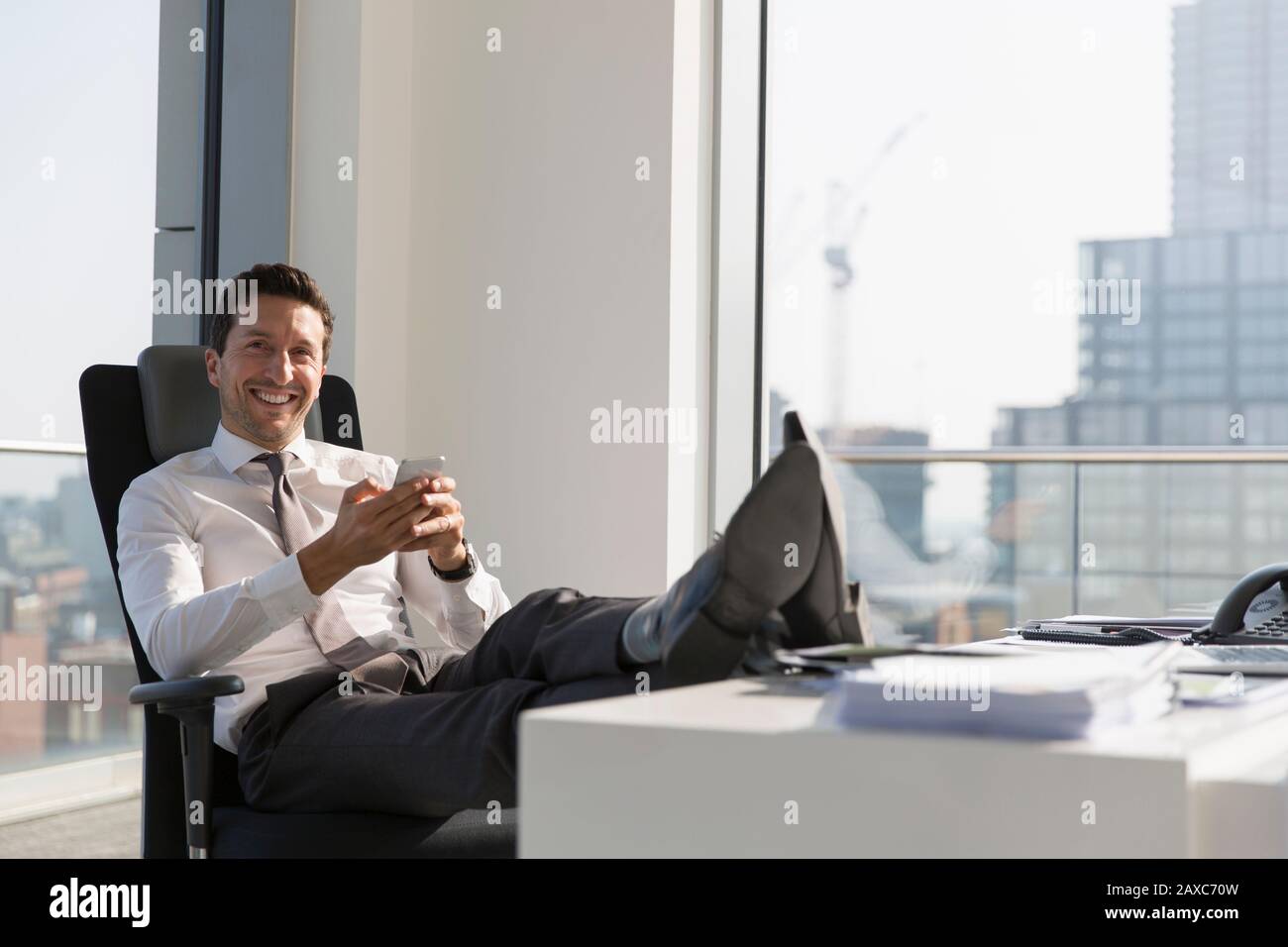 Hochformat selbstbewusster Geschäftsmann mit Smartphone mit Füßen auf dem Schreibtisch im Hochhaus Stockfoto