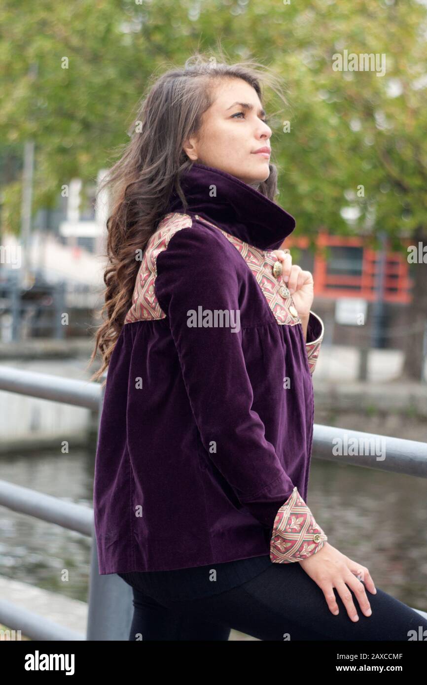 Kontemplative junge Frau in violettem Samtmantel, der am Zaun steht und den Fluss überblickt Stockfoto