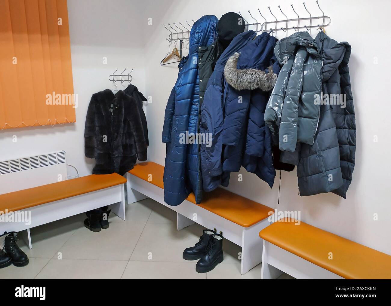 Jacken und Winteroberbekleidung hängen an Haken an der Wand. Im Umkleideraum der Frauen hängen Jacken, Fellmäntel und Mäntel. Orangefarbene Bänke, Schuhe und Klo Stockfoto