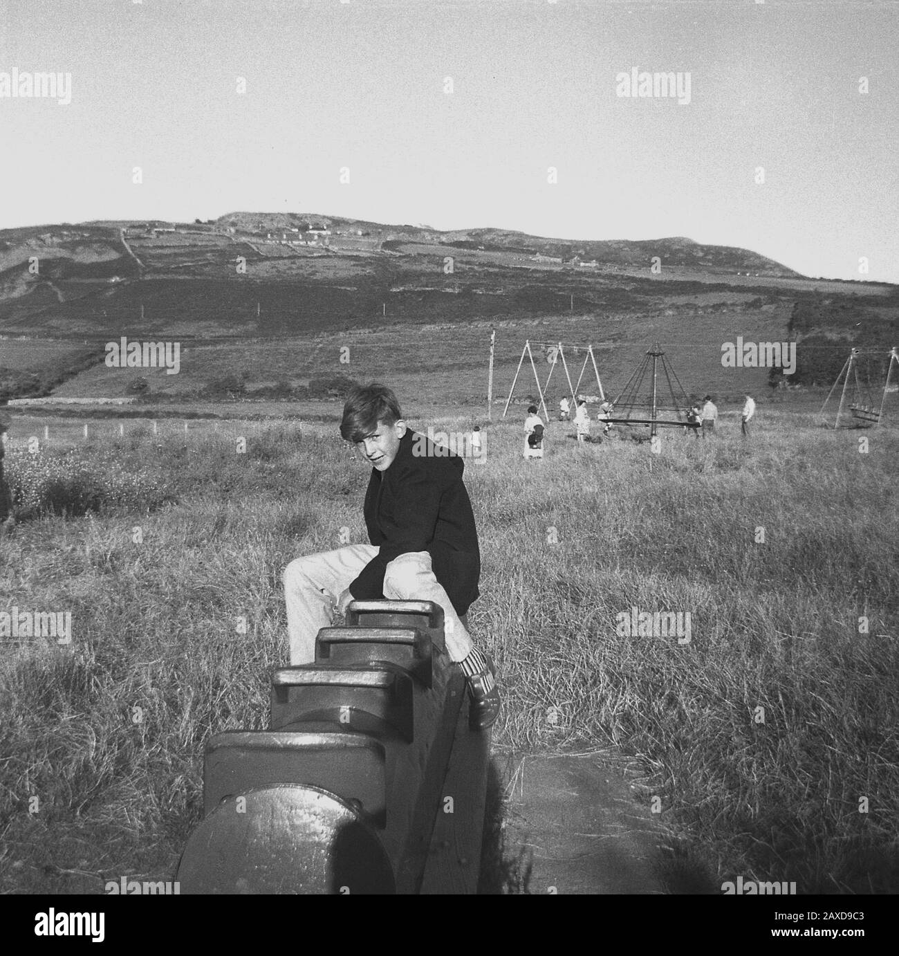 1950er Jahre, historisch gesehen, saß ein Teenager-Junge auf einem Metallschaukelpferd, möglicherweise einem britischen Wicksteed-Modell, draußen auf einem Spielplatz auf einem Grasfeld, in einem ländlichen Tal in der Nähe von Liverpool, England, Großbritannien. In der Ferne gesehen, spielen einige Kinder auf einem Hexenhut, ein häufiger Anblick auf Spielplätzen in dieser Zeit. Stockfoto