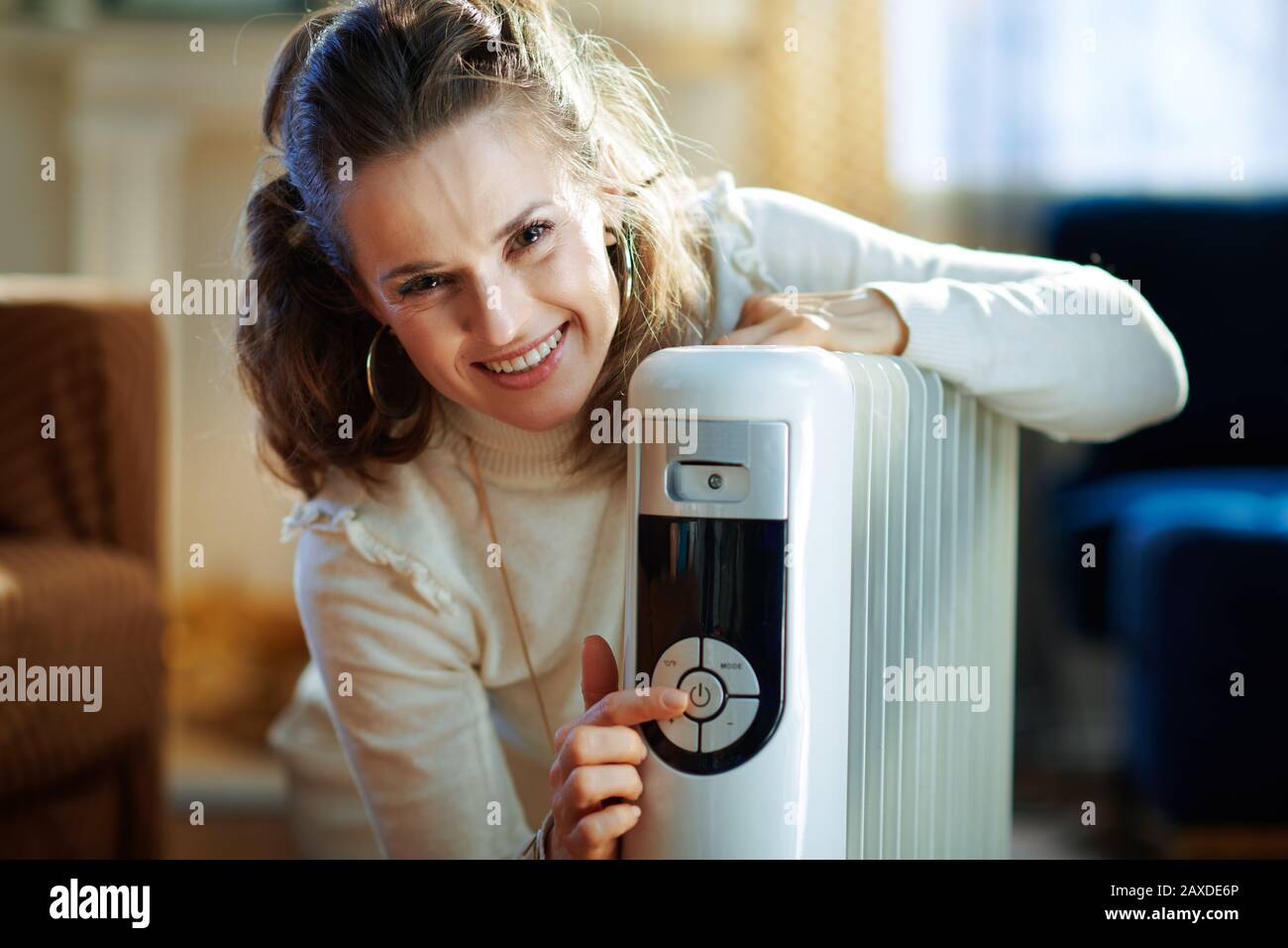 Fröhliche modische Frau im weißen Pullover und Rock im modernen Wohnzimmer im sonnigen Wintertag schalten Sie den weißen Elektro-Ölkühler ein. Stockfoto