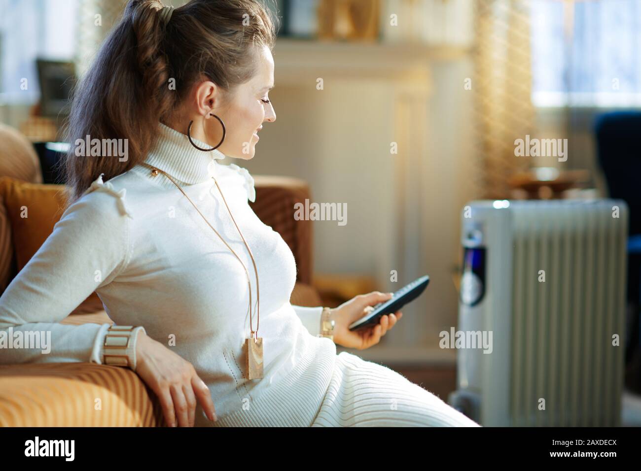 Lächelnde, moderne Frau in weißem Pullover und Rock im modernen Wohnzimmer am sonnigen Wintertag in der Nähe der Couch und des weißen Elektro-Ölkühlers Stockfoto