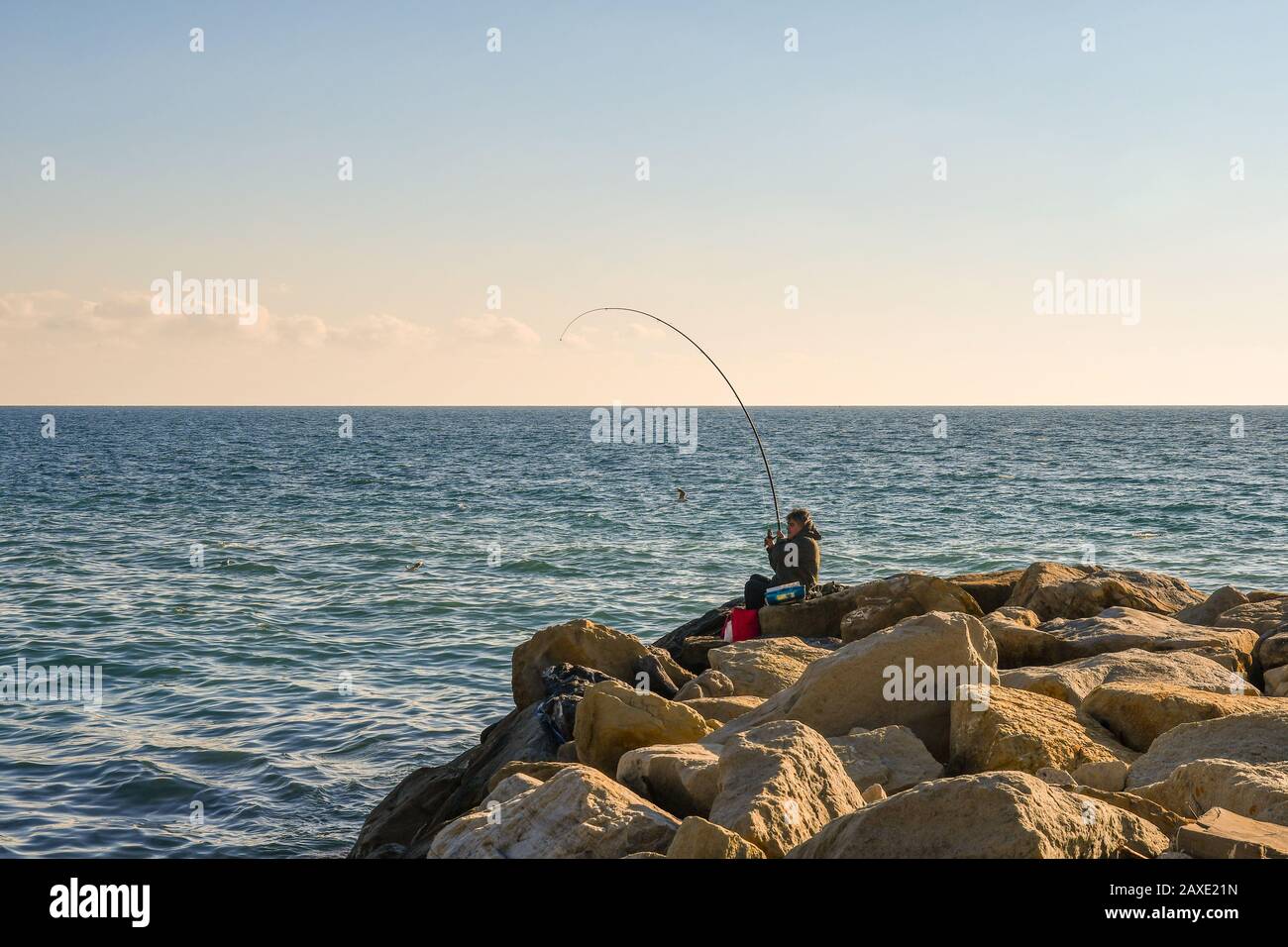 Ein Mann hat gerade einen Fisch mit einer Rute gefangen und an einem sonnigen Wintertag auf den Felsen eines Wellenbrechers angelt, Sanremo, Imperia, Ligurien, Italien Stockfoto