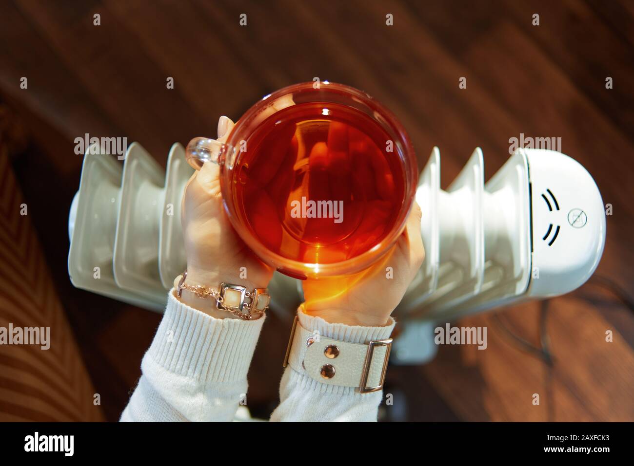 Die stilvolle 40-jährige Frau im modernen Haus im sonnigen Wintertag, die eine Tasse heißen Tee in der Nähe des weißen Elektro-Ölkühlers hält, ist ganz im Nahbereich. Stockfoto