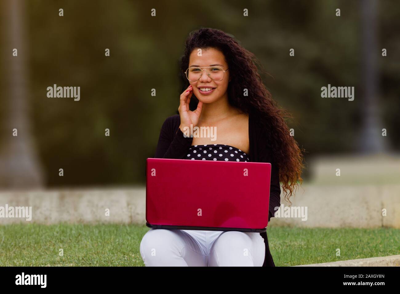 Afroamerikanische Frau, die an einem roten Laptop arbeitet. Attraktives hispanisches Mädchen mit Brille, die auf die Kamera blickt Stockfoto