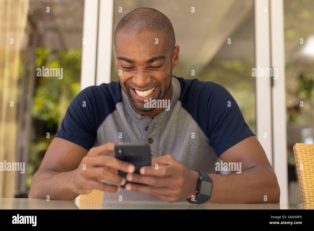 Vorderansicht Nahaufnahme eines afroamerikanischen Mannes mit grauem T-Shirt auf der Terrasse, mit seinem Handy und lächelnd Stockfoto