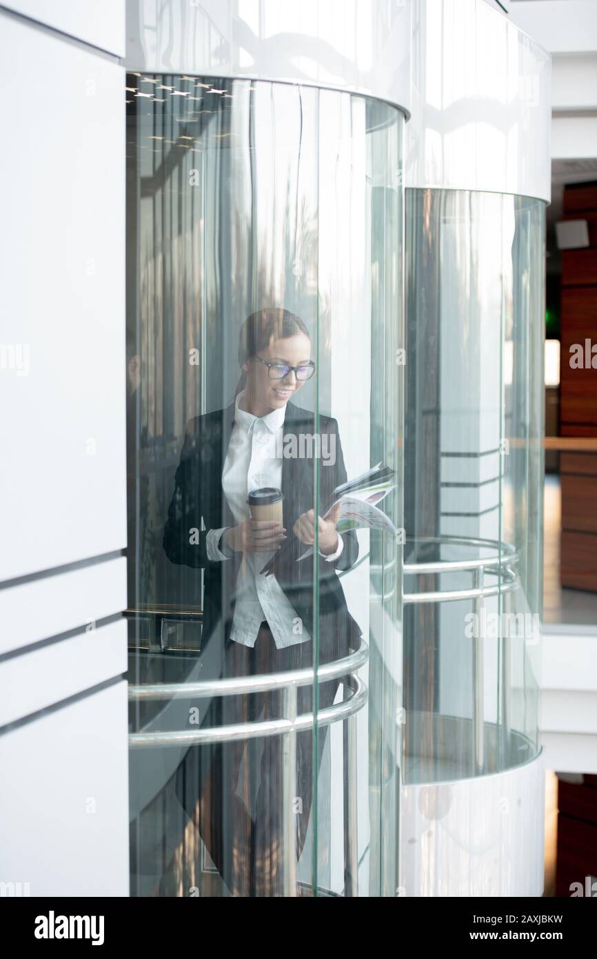 Lächelnde junge Geschäftsfrau in Brille, die Kaffee trinkt und im Aufzug des Bürogebäudes Papiere betrachtet Stockfoto