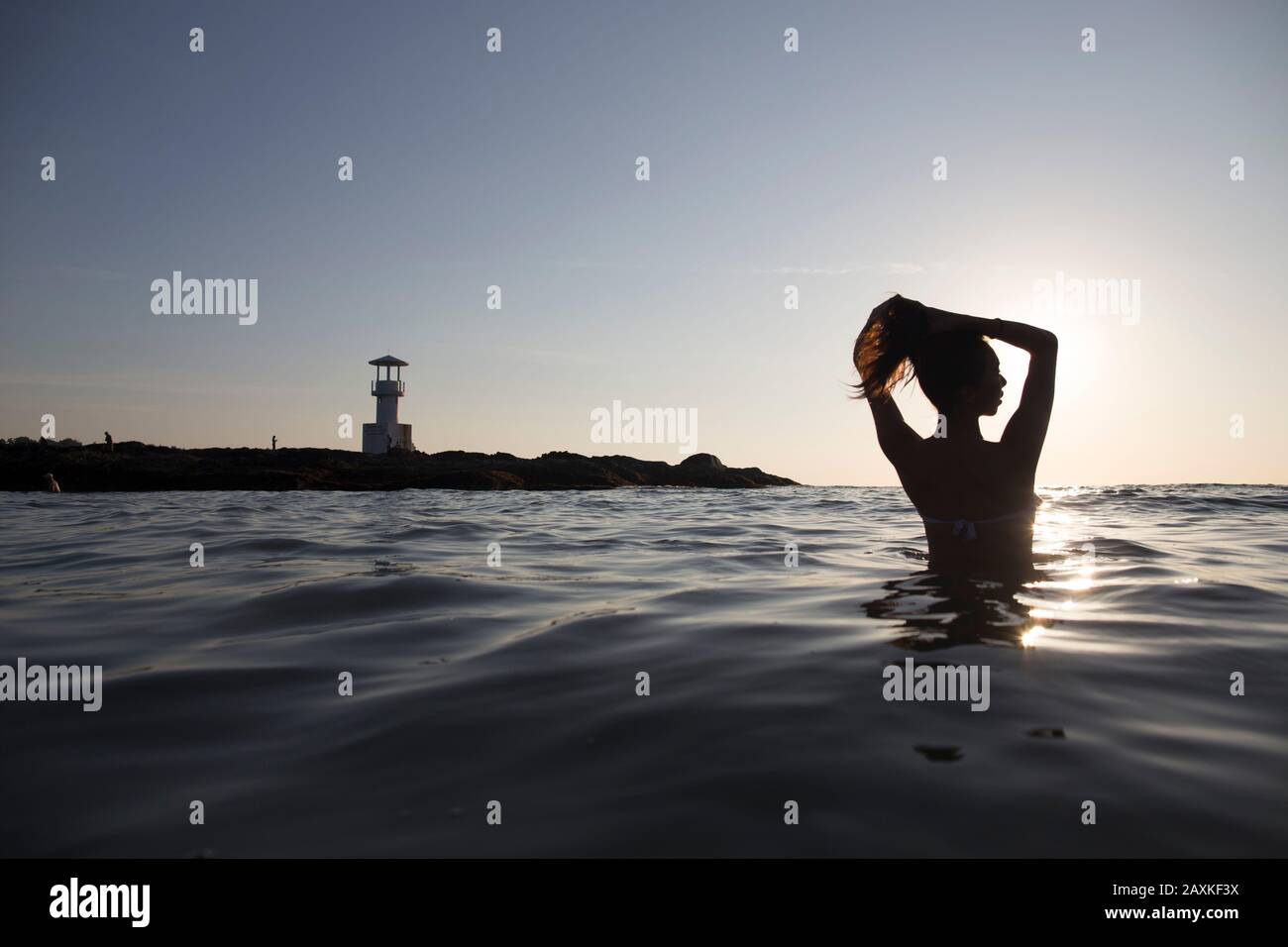 Rückansicht der Frau, die bei Sonnenuntergang im Meer baden kann, Leuchtturm in der Ferne. Stockfoto