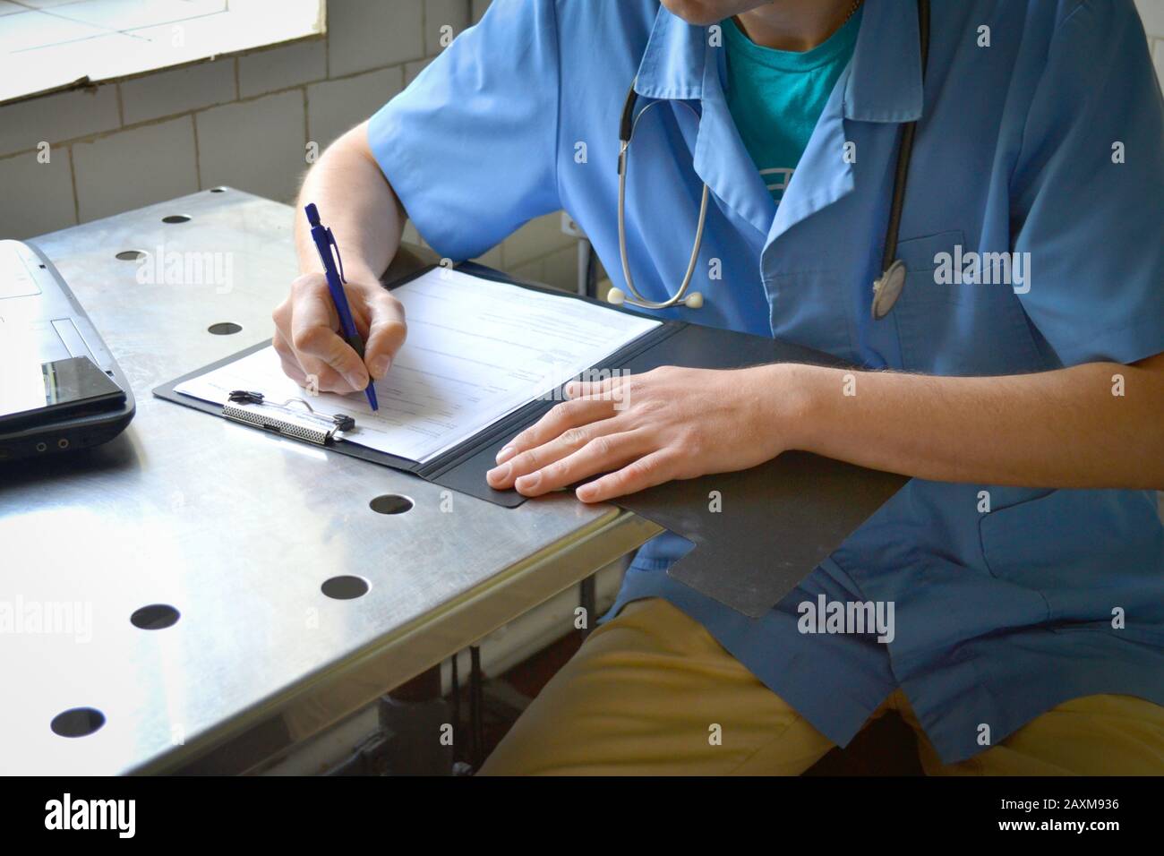 Doktor in blauer Uniform an einem Metalltisch sitzend und schreibt in einem Dokument Stockfoto